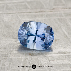 A 1.12-carat heated Ceylon sapphire, cushion-cut with facets, shown on a textured light gray background and "EARTH'S TREASURY" at the bottom.