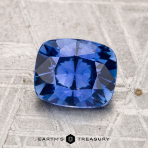 A 1.84-carat Ceylon sapphire gemstone with a cushion cut is displayed on a textured gray surface with "EARTH'S TREASURY" written below.