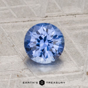 A 1.19-carat, heated Ceylon sapphire in a round brilliant cut is shown on a textured silvery surface with the "Earth's Treasury" logo at the bottom.