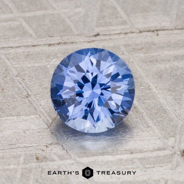 1.19-Carat Ceylon Sapphire (Heated) A 1.19-carat, heated Ceylon sapphire in a round brilliant cut is shown on a textured silvery surface with the "Earth's Treasury" logo at the bottom.
