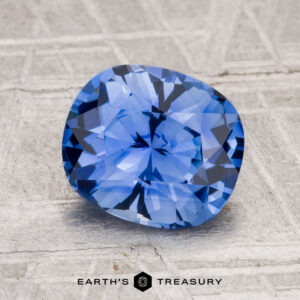 A 2.21-carat Ceylon sapphire with a cushion cut is displayed on a textured surface, with the text "EARTH'S TREASURY" shown below.