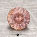 A round, faceted 3.04-carat Umba sapphire with a pink hue rests on a textured gray surface. The "Earth's Treasury" logo appears at the bottom of the image.