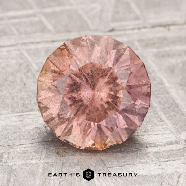 3.04-Carat Umba Sapphire A round, faceted 3.04-carat Umba sapphire with a pink hue rests on a textured gray surface. The "Earth's Treasury" logo appears at the bottom of the image.