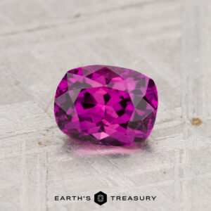 A 1.28-carat, cushion-cut Ceylon Sapphire (heated) sparkles on a textured metallic surface featuring the "Earth's Treasury" logo and text at the bottom.