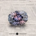 A 1.33-carat Montana sapphire, faceted in an oval shape with blue and pink hues, rests on a light-colored textured surface. "Earth's Treasury" is written at the bottom.