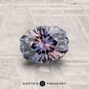 A 1.33-carat Montana sapphire, faceted in an oval shape with blue and pink hues, rests on a light-colored textured surface. "Earth's Treasury" is written at the bottom.