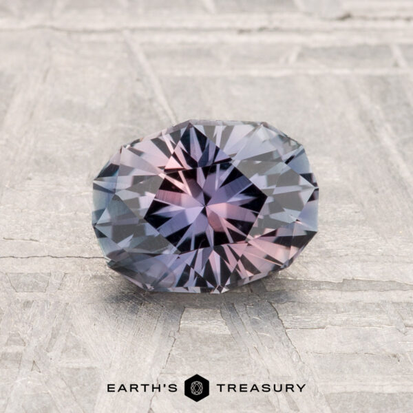 1.33-Carat Montana Sapphire A 1.33-carat Montana sapphire, faceted in an oval shape with blue and pink hues, rests on a light-colored textured surface. "Earth's Treasury" is written at the bottom.