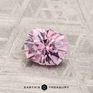 A faceted, light purple 0.92-carat Montana sapphire rests on a textured metallic surface with "EARTH'S TREASURY" displayed at the bottom.