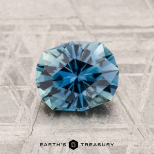 A 1.56-carat Montana sapphire, round brilliant-cut and blue, is displayed on a textured gray surface with "EARTH'S TREASURY" at the bottom.