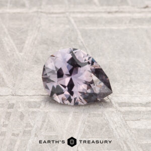 A 0.68-carat Montana sapphire, pear-shaped and faceted with a light purple hue, rests on a textured metallic surface. "EARTH'S TREASURY" appears at the bottom.