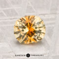 A round, faceted yellow-orange 1.44-carat Montana sapphire rests on a textured surface, with the label "Earth's Treasury" at the bottom.