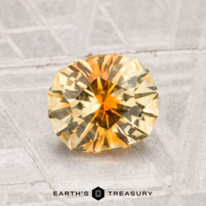 A round, faceted yellow-orange 1.44-carat Montana sapphire rests on a textured surface, with the label "Earth's Treasury" at the bottom.