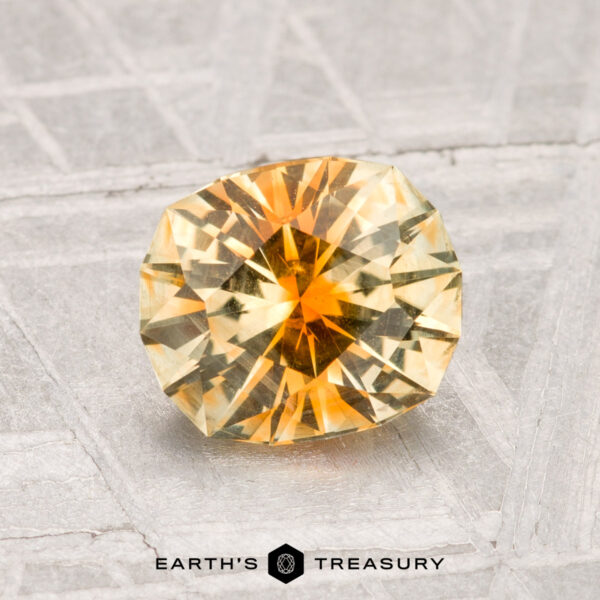 1.44-Carat Montana Sapphire A round, faceted yellow-orange 1.44-carat Montana sapphire rests on a textured surface, with the label "Earth's Treasury" at the bottom.