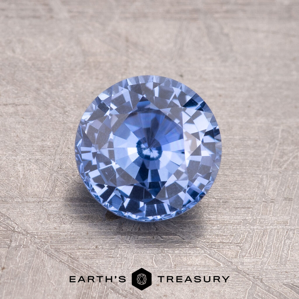 A 1.50-Carat Ceylon Sapphire in a round brilliant cut is displayed on a textured surface with the "EARTH'S TREASURY" logo and text below—ideal for anyone seeking an exceptional gemstone.