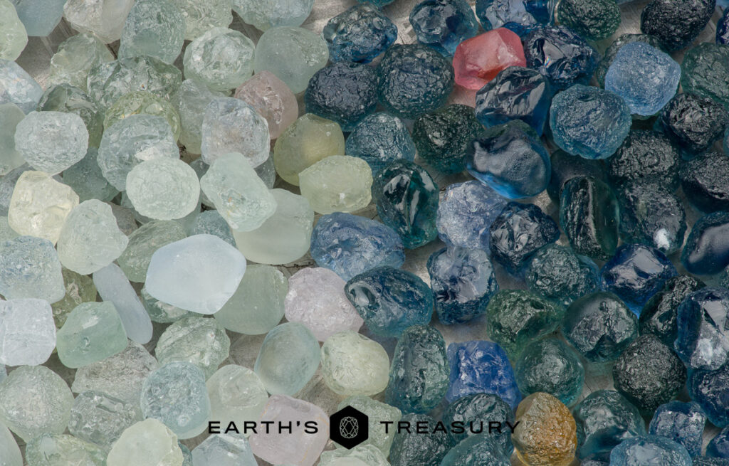 A collection of rough, uncut gemstones in various greens and blues, arranged side by side to showcase the natural beauty of no heat sapphires. "Earth's Treasury" logo appears below. Product: Stacked from 36 images. Method=B (R=14,S=2).