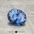 A striking 1.55-carat Ceylon sapphire, oval-cut and displaying vivid cornflower blue hues, lies on a textured gray surface with "EARTH'S TREASURY" printed at the bottom.