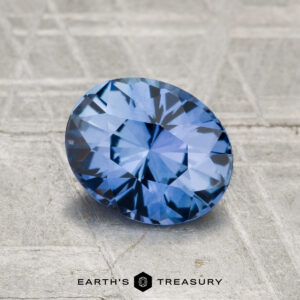 A striking 1.55-carat Ceylon sapphire, oval-cut and displaying vivid cornflower blue hues, lies on a textured gray surface with "EARTH'S TREASURY" printed at the bottom.