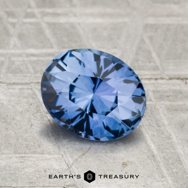 1.55-Carat Ceylon Sapphire A striking 1.55-carat Ceylon sapphire, oval-cut and displaying vivid cornflower blue hues, lies on a textured gray surface with "EARTH'S TREASURY" printed at the bottom.