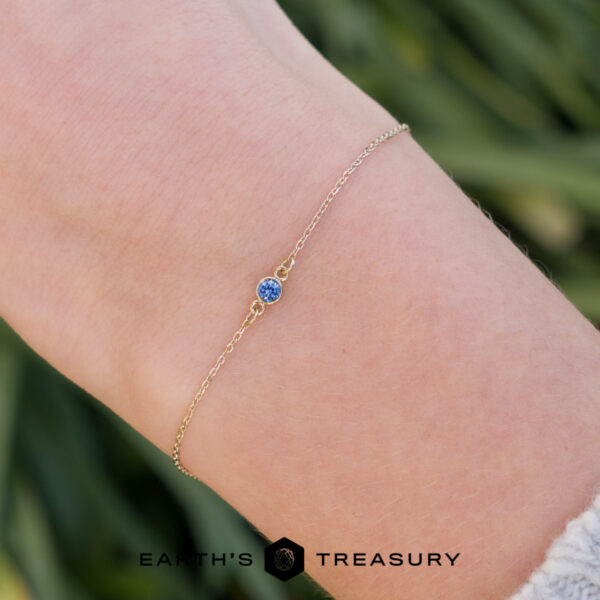 The "Lumi" Bracelet On Hand A wrist adorned with the _DSC1614-1200-BW-2 bracelet—a delicate gold chain featuring a small blue gemstone—stands out against a softly blurred, green-toned background.