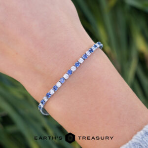 A close-up of a wrist wearing product _DSC1635-1200-BW, a silver tennis bracelet with alternating blue and clear stones, set against blurred green foliage in the background.