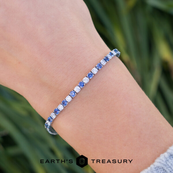 The "Adeline" Bracelet On Hand A close-up of a wrist wearing product _DSC1635-1200-BW, a silver tennis bracelet with alternating blue and clear stones, set against blurred green foliage in the background.