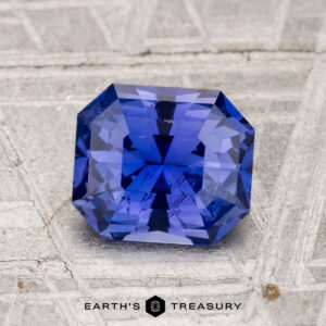A 1.63-carat Ceylon sapphire with an emerald cut is displayed on a textured gray surface, with "Earth's Treasury" shown below.