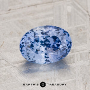 A 2.11-carat Ceylon sapphire with an oval cut and light sky blue hue rests on a textured surface, with "EARTH'S TREASURY" at the bottom.