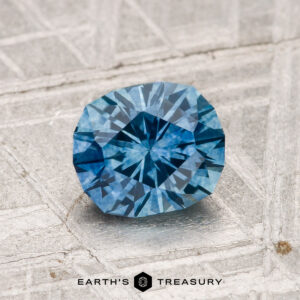 A 1.32-carat heated Montana Sapphire with a geometric facet pattern is displayed on a textured surface, with "EARTH'S TREASURY" beneath it.