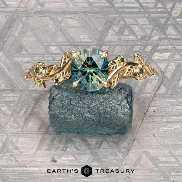 The Deluxe "Ivey" ring in 14K yellow gold with 1.58-carat Australian sapphire The Deluxe "Ivey" ring in 14k yellow gold with a 1.58-carat Australian sapphire is shown atop a rough blue-green stone, with the "Earth's Treasury" logo below.