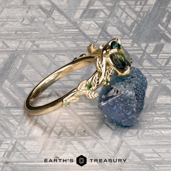 The Deluxe "Ivey" ring in 14K yellow gold with 1.58-carat Australian sapphire The Deluxe "Ivey" ring in 14k yellow gold, featuring leaf designs and a 1.58-carat Australian sapphire, rests against a rough blue stone on a geometric patterned surface.