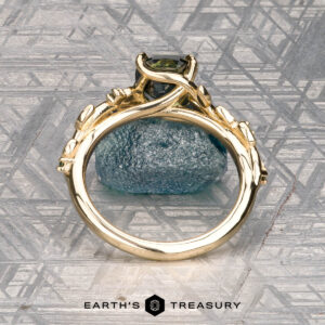 The Deluxe "Ivey" ring in 14k yellow gold with a 1.58-carat Australian sapphire stands upright against a blue textured surface, displayed before a geometric patterned background with the Earth's Treasury logo below.