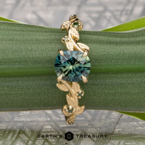 The Deluxe "Ivey" ring in 14k yellow gold with a 1.58-carat Australian sapphire, featuring an intricate leaf and vine design, is displayed on a green plant stem. Below it appears the text "EARTH'S TREASURY.