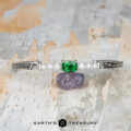 The "Amaya" Bangle in 14k white gold, featuring a 3.03-carat Tsavorite garnet and diamonds, rests on a purple stone atop a textured surface. The text "EARTH'S TREASURY" appears at the bottom.