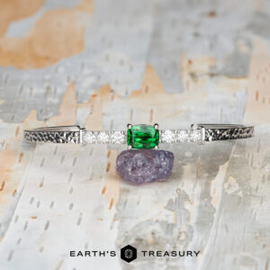 The "Amaya" Bangle in 14k white gold, featuring a 3.03-carat Tsavorite garnet and diamonds, rests on a purple stone atop a textured surface. The text "EARTH'S TREASURY" appears at the bottom.