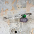The "Amaya" Bangle in 14k white gold, featuring a 3.03-carat Tsavorite garnet and diamonds, rests on a purple stone atop a textured surface. The text "EARTH'S TREASURY" appears at the bottom.