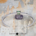 The "Amaya" Bangle in 14k white gold, featuring a 3.03-carat Tsavorite garnet and diamonds, rests on a purple stone atop a textured surface. The text "EARTH'S TREASURY" appears at the bottom.