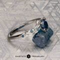 The "Aralia" ring in platinum, featuring a 1.03-carat Montana sapphire and leaf details, is shown beside a rough blue stone on a textured surface. The Earth's Treasury logo appears at the bottom.