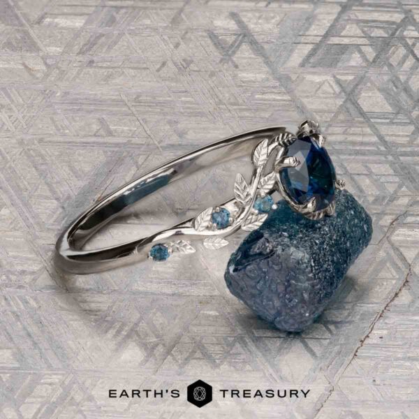 The "Aralia" ring in platinum with 1.03-carat Montana sapphire The "Aralia" ring in platinum, featuring a 1.03-carat Montana sapphire and leaf details, is shown beside a rough blue stone on a textured surface. The Earth's Treasury logo appears at the bottom.