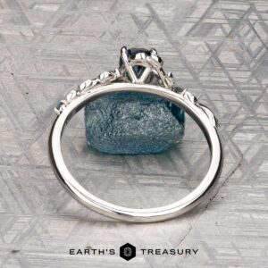 The "Aralia" ring in platinum with a 1.03-carat Montana sapphire stands upright, displaying its band and setting, with a rough blue stone behind it on a patterned surface. The Earth's Treasury logo appears below.
