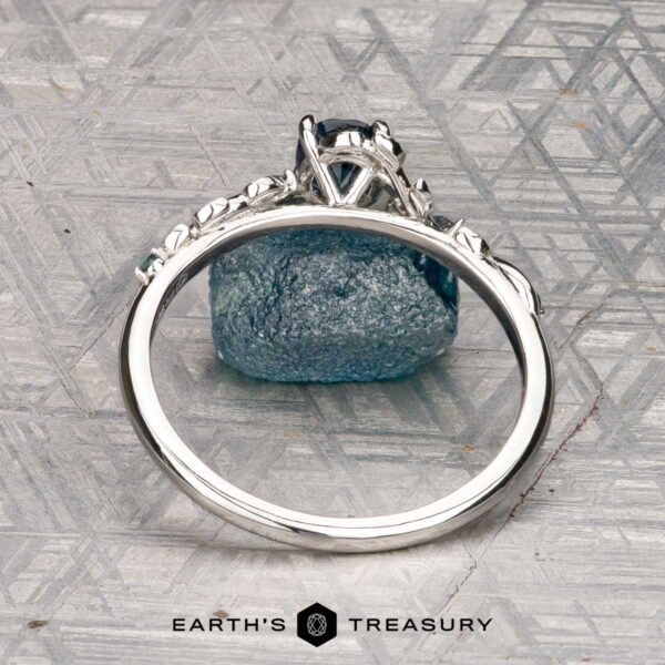 The "Aralia" ring in platinum with 1.03-carat Montana sapphire The "Aralia" ring in platinum with a 1.03-carat Montana sapphire stands upright, displaying its band and setting, with a rough blue stone behind it on a patterned surface. The Earth's Treasury logo appears below.