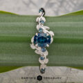 The "Aralia" ring in platinum, featuring a 1.03-carat Montana sapphire and leaf details, is shown on a green plant stem with the Earth’s Treasury logo below.