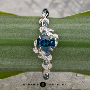 The "Aralia" ring in platinum, featuring a 1.03-carat Montana sapphire and leaf details, is shown on a green plant stem with the Earth’s Treasury logo below.