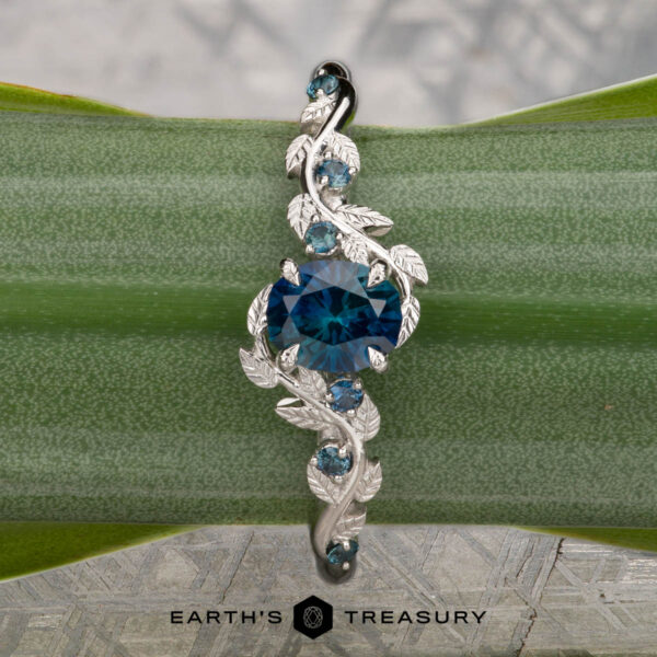 The "Aralia" ring in platinum with 1.03-carat Montana sapphire The "Aralia" ring in platinum, featuring a 1.03-carat Montana sapphire and leaf details, is shown on a green plant stem with the Earth’s Treasury logo below.