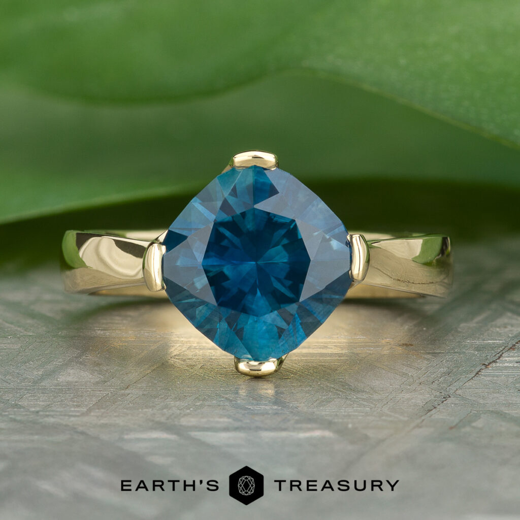 The "Stacked from 15 images. Method=B (R=14,S=2)" gold ring showcases a square-cut blue gemstone at its center, displayed on a reflective surface with green leaves in the background.