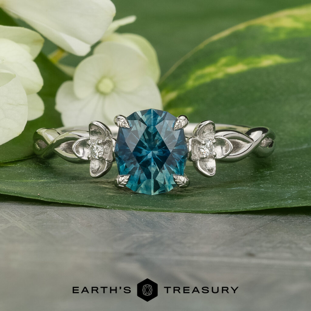 A silver ring with a blue oval gemstone and two small clear stones is displayed on a green leaf with white flowers. The Earth's Treasury logo is at the bottom. Product: Stacked from 17 images. Method=B (R=14,S=2).