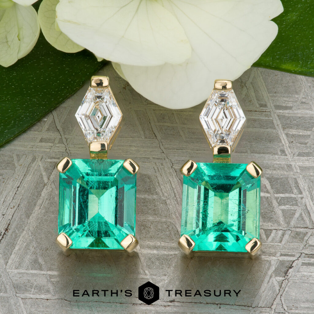 Stacked from 28 images. Method=B (R=14,S=2): Emerald-cut green gemstones set in gold with geometric diamond accents, shown on a textured surface with white flowers and green leaves in the background.