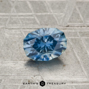 A round, faceted 1.03-carat heated Montana sapphire with a blue hue rests on a textured metallic surface, with "EARTH'S TREASURY" displayed below.