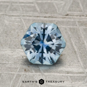 A 1.29-carat heated Montana sapphire, light blue and round brilliant-cut with an octagonal outline, is displayed on a textured gray surface. "EARTH'S TREASURY" appears at the bottom.