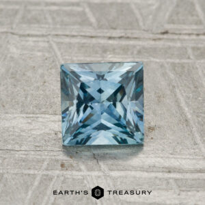 A 0.90-carat heated Montana sapphire, square-cut and blue, displayed on a textured gray surface with "EARTH'S TREASURY" at the bottom.