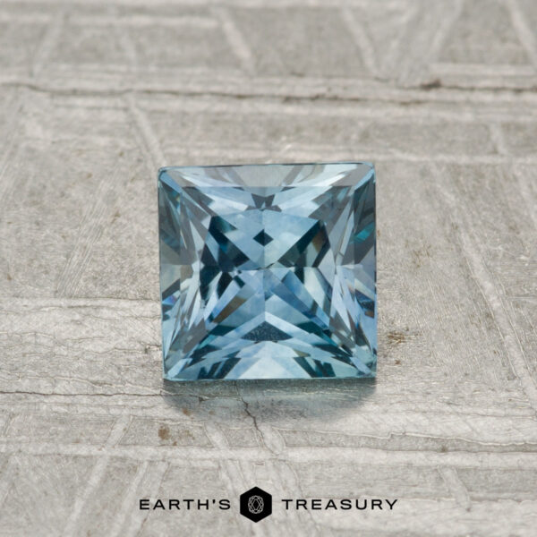 0.90 -Carat Montana Sapphire (Heated) A 0.90-carat heated Montana sapphire, square-cut and blue, displayed on a textured gray surface with "EARTH'S TREASURY" at the bottom.